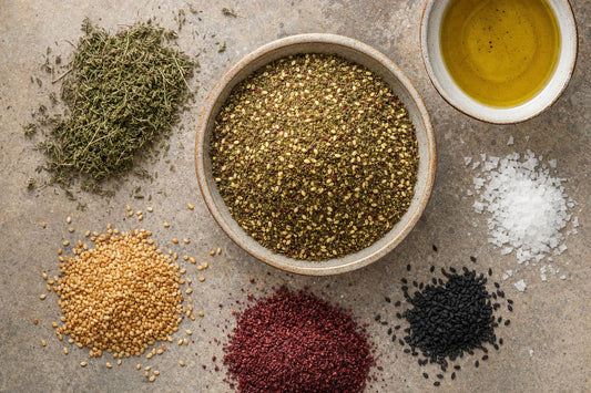 Zatar Gewürzmischung in einer Schale, daneben die Zutaten: wilder Thymian, gerösteter Sesam, roter Sumach, Schwarzkümmel, Salzflocken und Olivenöl