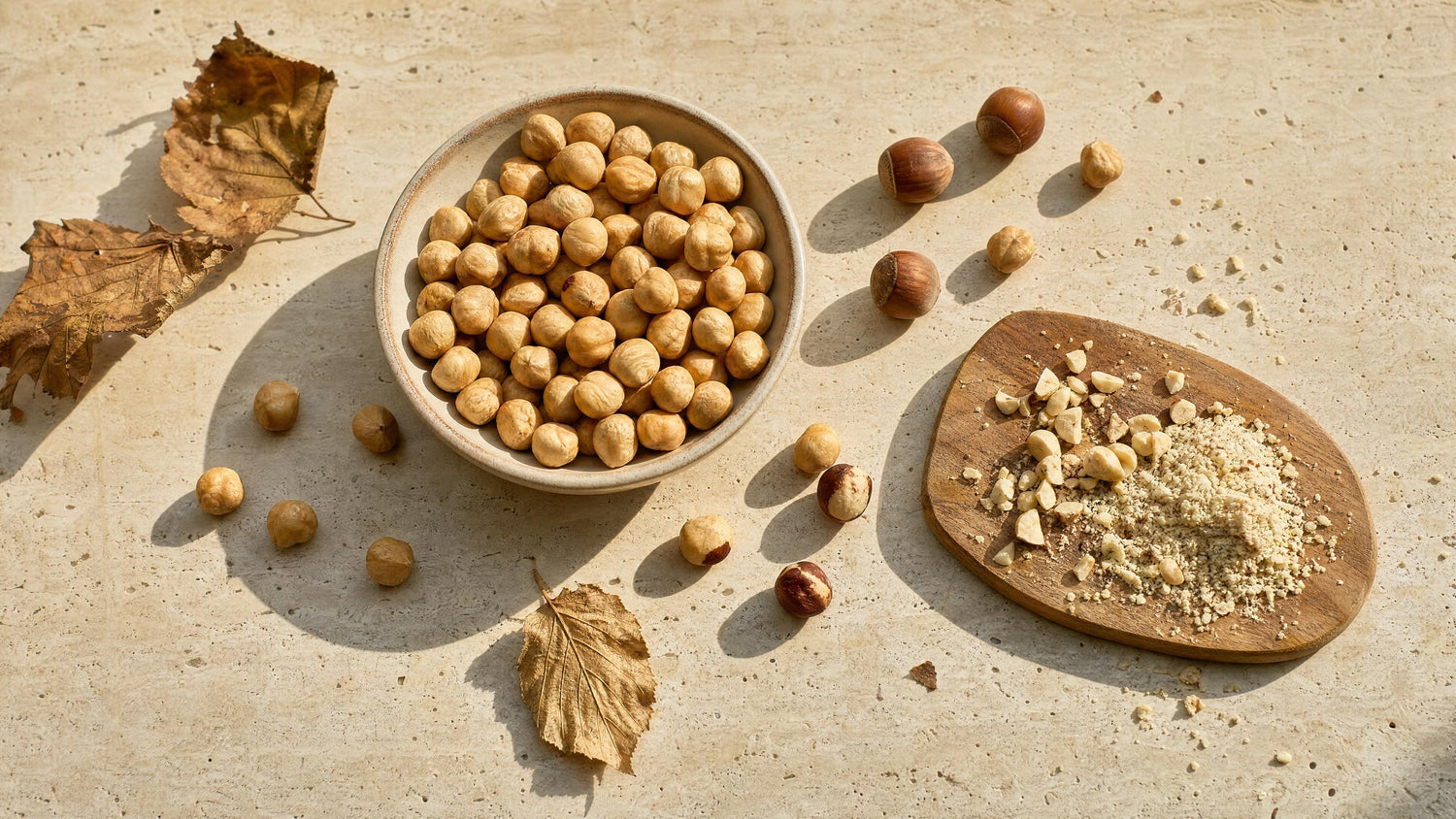 Geröstete Piemonteser Haselnüsse in einer Schale auf Travertin, daneben Nüsse in der Schale und gehackte Haselnüsse auf einem Holzbrett