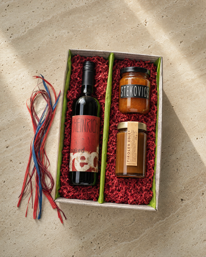 Österreichisches Gourmet-Präsent — Heinrich naked red, Stekovics Marille & Tiroler Edelkaramell im offenen 2er-Karton, Top-Down Editorial auf Travertin mit rotem Sizzlepack
